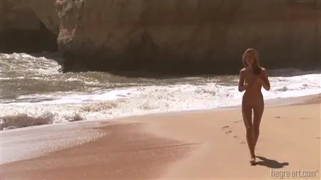Обнаженная блондинка резвится на пляже и загорает на горячем песке